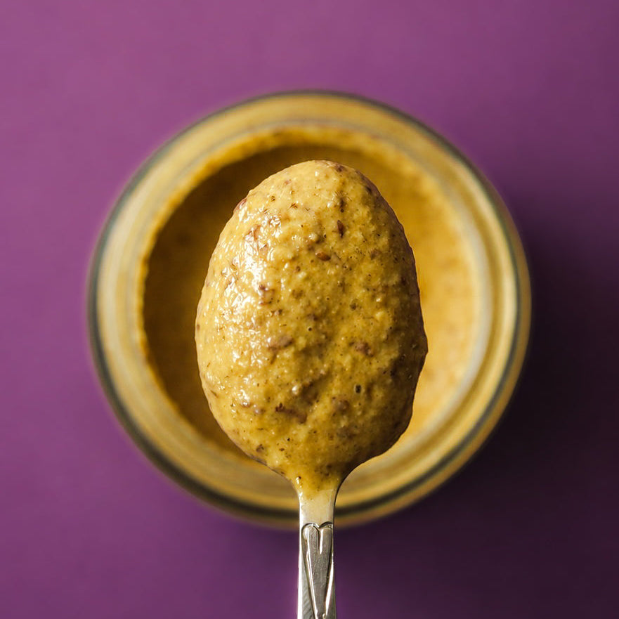 A spoonful of thick, textured super seed spread held above an open glass jar against a solid purple backgro